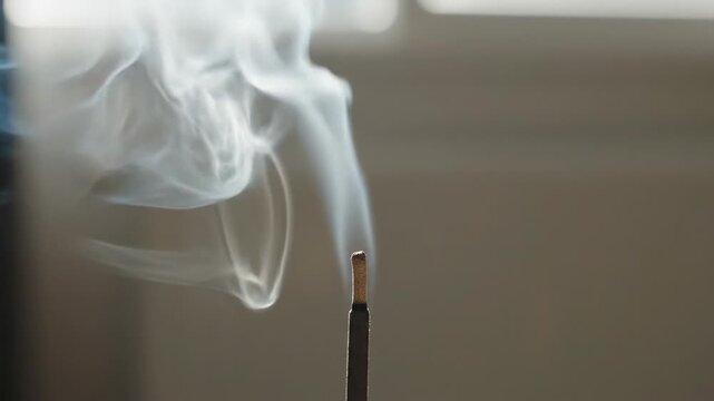 Wisps of White Incense Smoke Curl and Rise from a Dark Stick Against a Softly Blurred Neutral Background in Natural Light