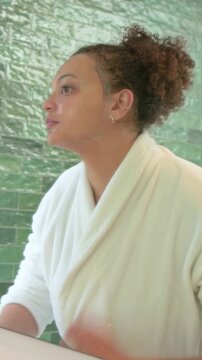 Vertical video: Leaning woman in white bathrobe rinsing face at bathroom sink, grabbing towel