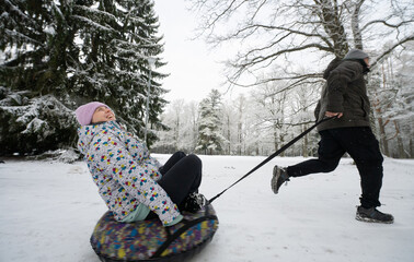 Man runs and pulls an inflatable donut sled with a girl in the snow in the winter park