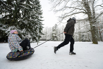 Man runs and pulls an inflatable donut sled with a girl in the snow in the winter park