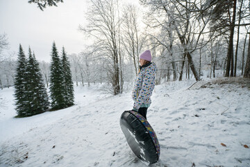 Girl stands and holds an inflatable donut sled in a winter park and looks at a snow slide