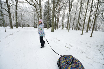 Girl pulling inflatable donut sled through a scenic snowy winter park.