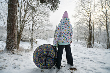 Girl stands and holds an inflatable donut sled in a winter park and looks at a snow slide
