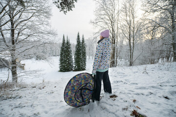 Girl stands and holds an inflatable donut sled in a winter park and looks at a snow slide