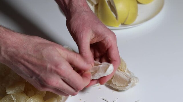 Close-up of hands peeling a grapefruit segment, meticulously removing the white pith to prepare a healthy and tasty snack on a clean white surface indoors.