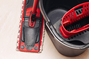 Floor cleaning mop and bucket set on beige surface. Red flat mop head and plastic bucket with wringer. Housekeeping equipment closeup for home cleaning routine use indoor chores concept.