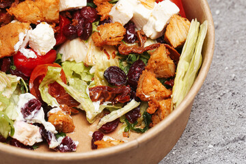 Fresh chicken salad bowl with lettuce cheese cranberries and croutons. Mixed greens with grilled chicken pieces and creamy dressing. Healthy lunch meal in eco paper bowl on table closeup