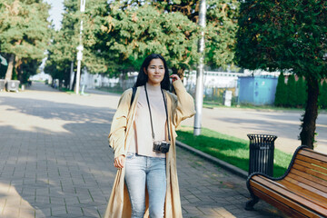 Obraz premium Woman in a long coat strolling along a park street with a camera, casual fashion, outdoor ambience, sunlight, urban lifestyle moment, relaxed pose and pedestrian background.
