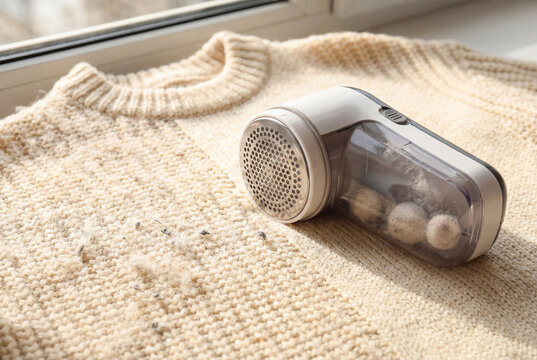 Electric fabric shaver or lint remover removing fuzz balls from beige wool sweater on windowsill