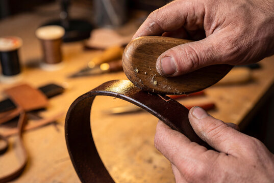 Leather craftsman burnishing edge of handmade brown leather belt with wooden slicker tool