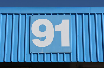Blue rectangular sign with the number 91 mounted on a corrugated metal wall