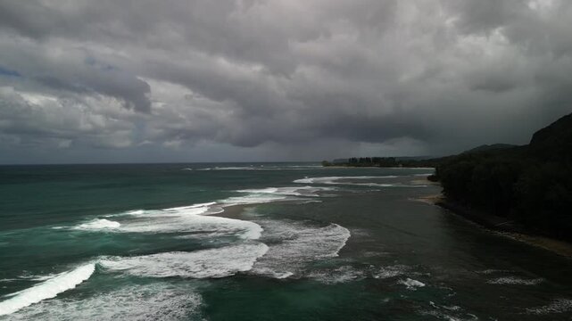 Moody aerial drone view of layered ocean waves rolling over shallow reef along the rugged Kauai coastline under dramatic storm clouds near Haena State Park
