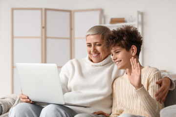 Fototapeta premium Teenage boy and his mother in warm sweaters with laptop video chatting on sofa at home