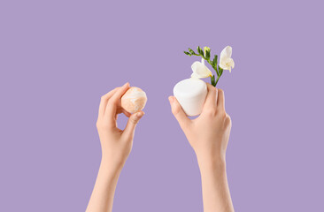 Female hands with bath bomb, flower and jar of cosmetic product on lilac background