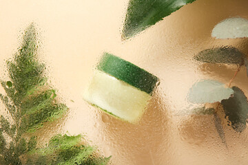 Cosmetic jar and green leaves behind wet glass on beige background