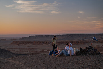 Happy friends with a young child relax on a rise at desert sunset during golden hour. The archaeological canyon of Khermen Tsav forms the dramatic backdrop in Mongolia.