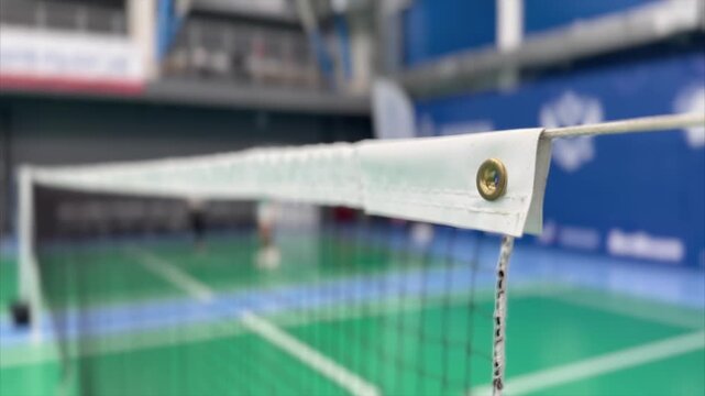 Video of a badminton net in sharp focus with an indoor court and athletes blurred in the background, creating depth and training atmosphere