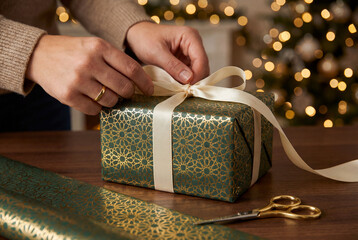 Person wrapping a gift box with elegant green and gold patterned paper and cream ribbon