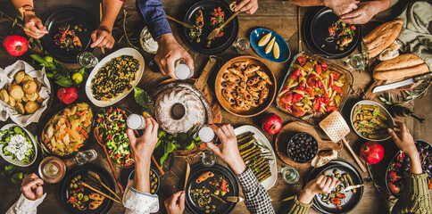 Lively friends and family dinner gathering around rustic wooden table sharing diverse dishes,...