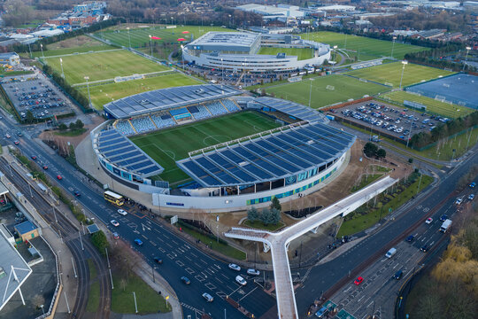 Aerial image of the Etihad Campus in Manchester at sunset, featuring the Joie Stadium and the pedestrian footbridge spanning Alan Turing Way