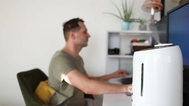 Modern air humidifier device during sitting workplace, young man using computer work on internet, enjoying aromatherapy steam scent from essential oil diffuser in room at home office.