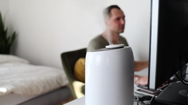 Modern air humidifier device during sitting workplace, young man using computer work on internet, enjoying aromatherapy steam scent from essential oil diffuser in room at home office.