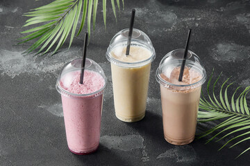 Milkshakes to go in plastic cups with dome lids and straws on dark stone background with tropical leaves, takeaway beverage menu concept