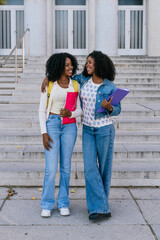 Fototapeta premium Two smiling young women, friends and students, embracing while walking down university campus stairs carrying books and folders