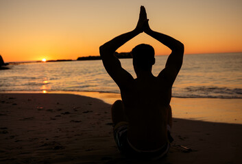 Obraz premium Dark silhouette is sitting cross-legged and meditating on sandy beach at sunset, sandals nearby