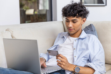 Fototapeta premium Man holding credit card typing on laptop, sitting on sofa in living room