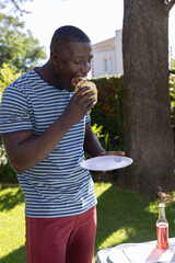 Fototapeta premium African american man standing on lawn eating hamburger from paper plate by table in backyard