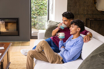 Naklejka premium Male friends sitting on sofa in living room holding ceramic mugs by fireplace