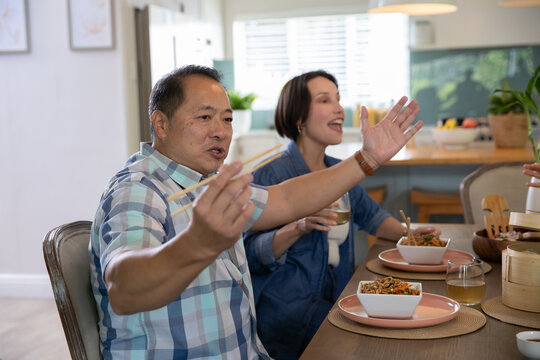 Asian couple sitting at dining table in modern kitchen using chopsticks and eating noodles - Powered by Adobe