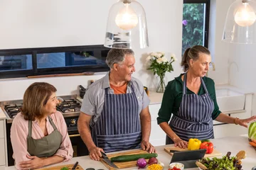 Selbstklebende Fototapeten Zu Kochen Senior friends wearing aprons preparing vegetables around kitchen island with tablet device  © wavebreak3