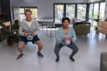 Diverse workout partners squatting with kettlebells in modern living room by glass doors