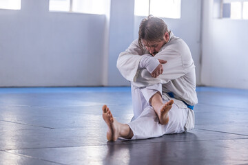 Martial artist in white gi, blue belt sitting and hugging knee on dojo mat near windows