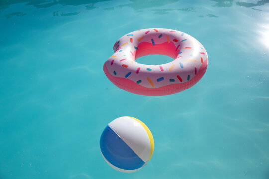 Pink donut float hovering on pool with striped ball under reflecting sunlight