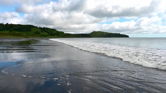 Clear skies meet rolling waves on the tranquil beach of Kunashir Island. Nature's beauty unfolds as the tides ebb and flow, painting a peaceful picture against the lush green backdrop.