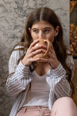 Naklejka premium Woman in her twenties sitting against marble wall holding beige mug, gazing left