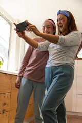 Asian mother and daughter holding smartphone in kitchen wearing headbands, under eye patches