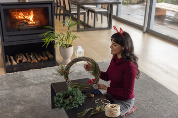 Naklejka premium Asian woman assembling twig wreath with pine cones, twine on coffee table in living room
