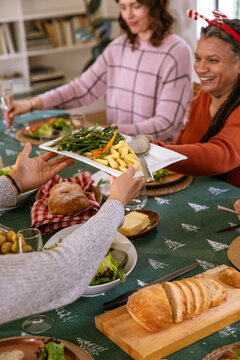 Diverse family in festive sweaters passing platter of green beans, potatoes around dining table