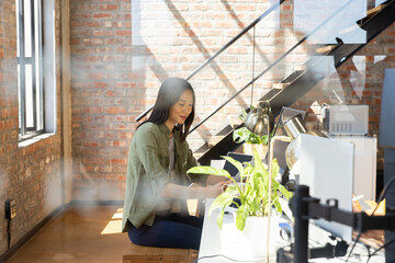 Asian woman working at loft-style office desk using laptop, dual monitors, desk lamp and plants