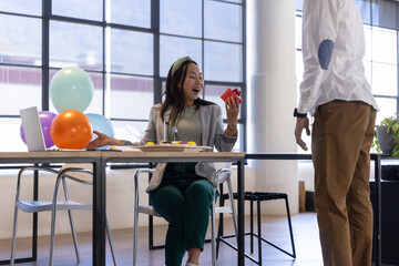 Obraz premium Diverse coworkers celebrating in office chinese woman holding red gift box by cake, balloons