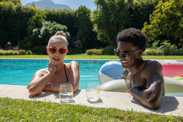 Diverse couple leaning on edge at home pool in swimwear holding pool tube, lemon-slice glasses