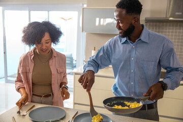 Diverse couple holding frying pan and spatula, cooking scrambled eggs on countertop in kitchen