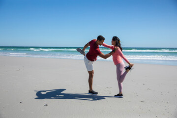 Diverse couple performing quad stretch on sandy beach by sea waves in sportswear and sneakers