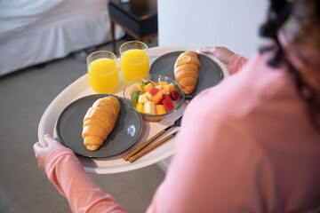 Adult woman with curly hair with white tray on bed with croissants, fruit bowl, orange juice