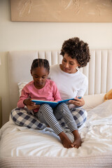 African american mother with daughter reading blue book on duvet-covered bed with teddy bear