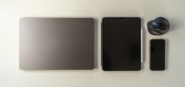 Clean flat lay of contemporary tech gadgets including laptop computer, tablet with stylus, smartphone, and mouse. Office workspace concept.